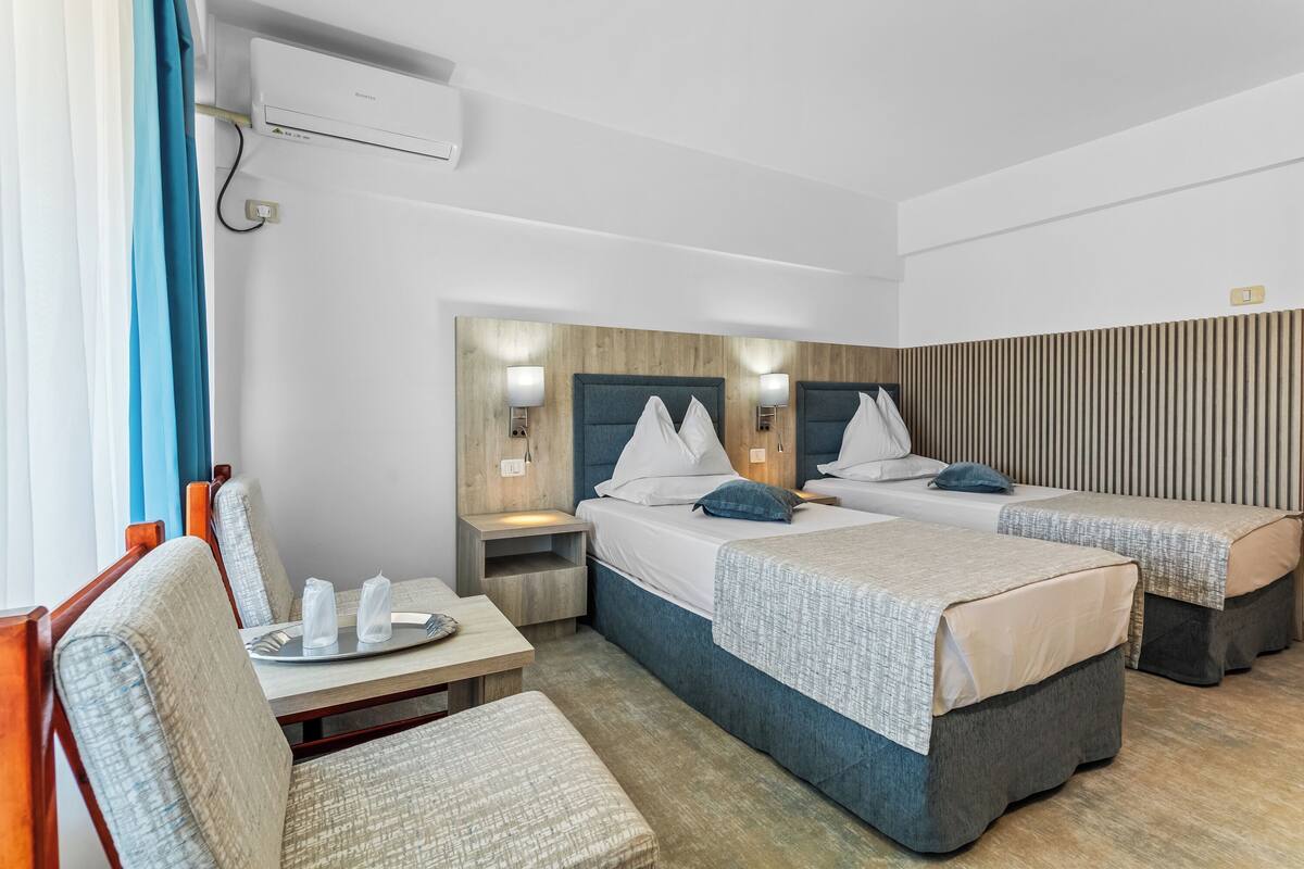 Une chambre d’hôtel avec deux lits, un bureau et une chaise. On y trouve une table de chevet avec une lampe, un climatiseur fixé au mur et une prise de courant.