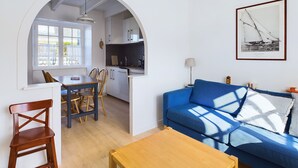 Een moderne woonkamer met een blauwe bank, een houten eettafel en stoelen. Er is een keuken met witte kasten en een hanglamp.