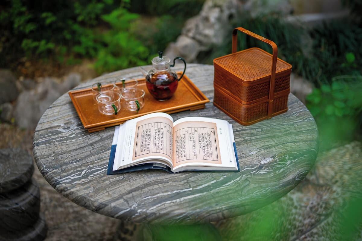 Un tavolo con un libro aperto, un teiera e un vassoio con bicchieri e un cesto.