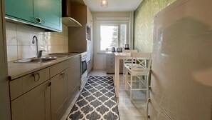 A compact kitchen with teal cabinets, a stainless steel sink, a built-in oven, and a patterned rug leading to a dining area with a white table and chairs. - Kotimaailma KotikolmioOikokatu (Lahti)