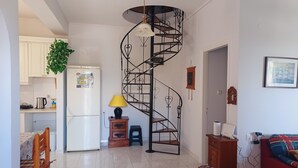 A compact living space with a spiral staircase, a dining area with a table and chairs, a kitchen with a refrigerator and cabinets, and a red sofa in the living area. - Villa Mariza at Aiantio (Salamina)