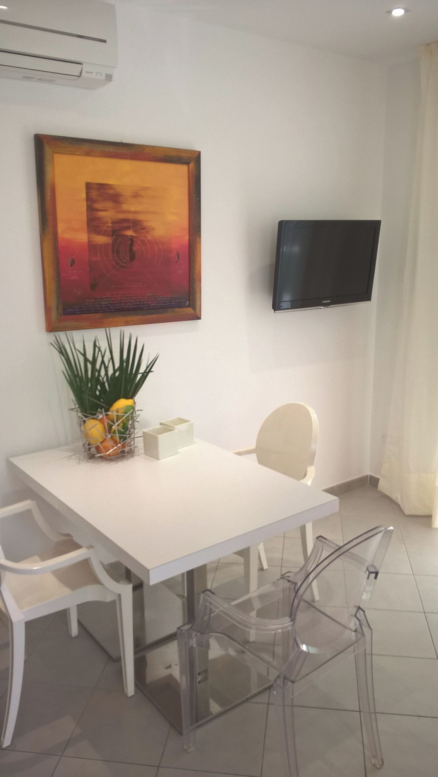 A modern dining area with a white table, clear chairs, a potted plant, and a flat-screen TV mounted on the wall.