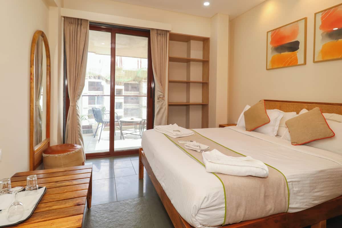 A hotel room with a large bed, a wooden side table with glasses, a mirror, and a sliding glass door leading to an outdoor area.