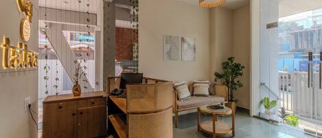 A modern living room with a sofa, coffee table, and a wicker chair. There is a desk with a computer and a telephone. A large window provides a view of the outside.