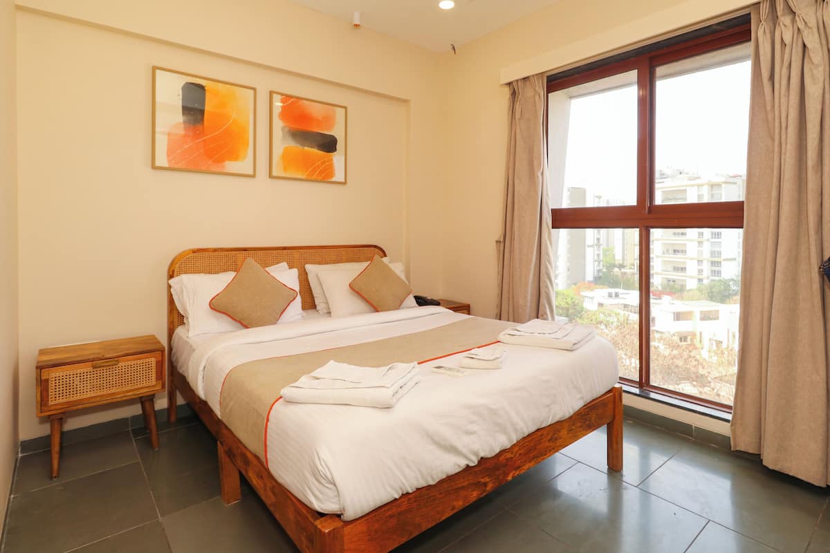 A neatly made bed with white linens and beige accents, a wooden headboard, a small wooden nightstand, and a large window with curtains.