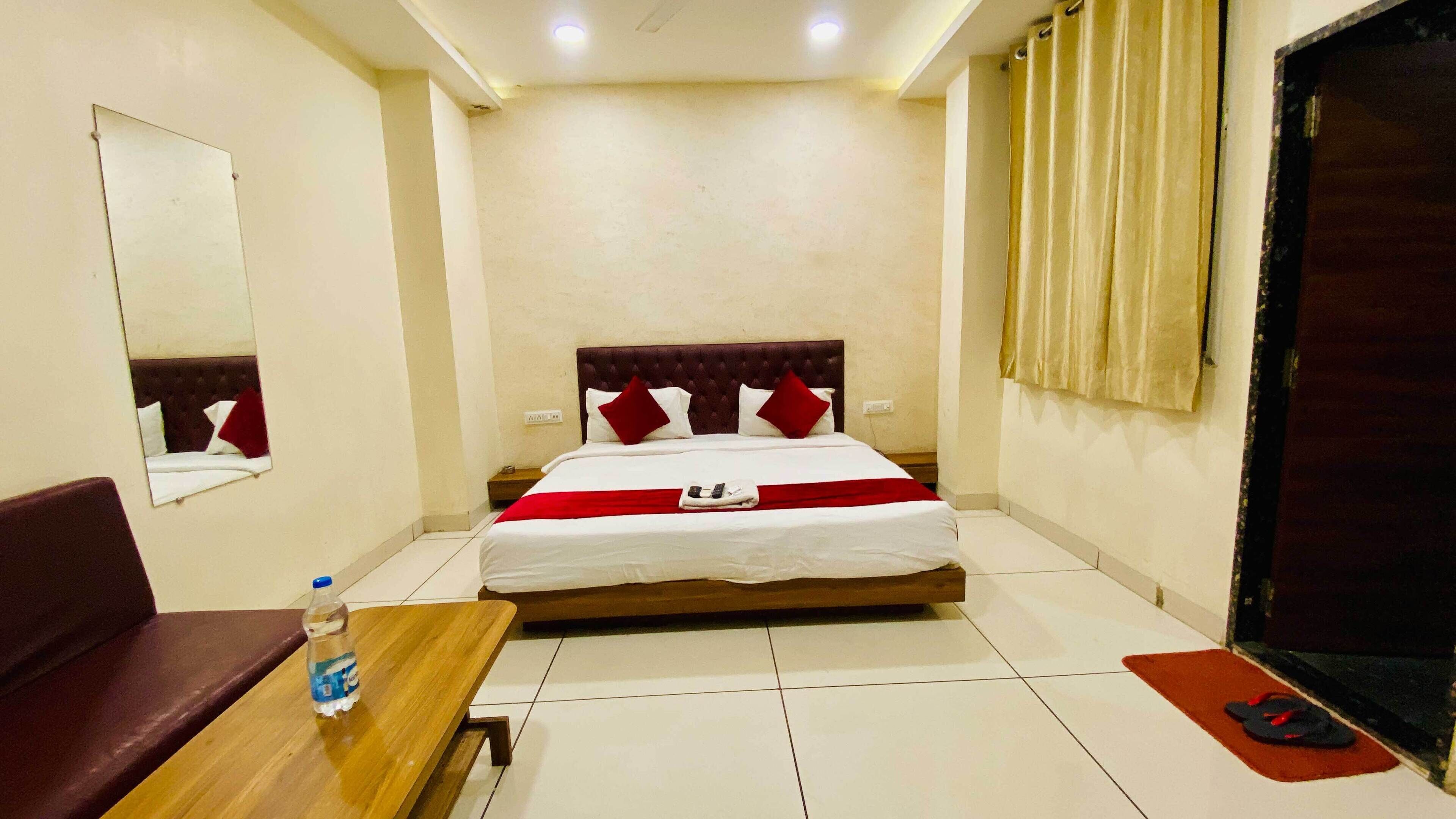 A hotel room with a bed, a wooden table with a water bottle, a red and white bedspread, a red and white pillow, a mirror on the wall, and a door with curtains.