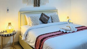 A neatly made bed with white linens and a pink throw blanket, a wall-mounted macrame piece, and a small table with a woven lamp beside the bed. - Alfa Kuta (Kuta)