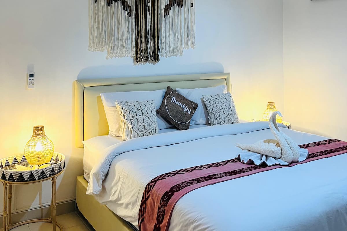 Un letto rifatto con lenzuola bianche e una coperta rosa, un pannello macramè a parete e un comodino con una lampada intrecciata accanto al letto.