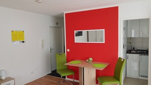 Een compact woongedeelte met een eethoek, bestaande uit een houten tafel en twee groene stoelen, een rode accentmuur, een kleine keuken met witte kastjes en een deur naar een andere kamer.