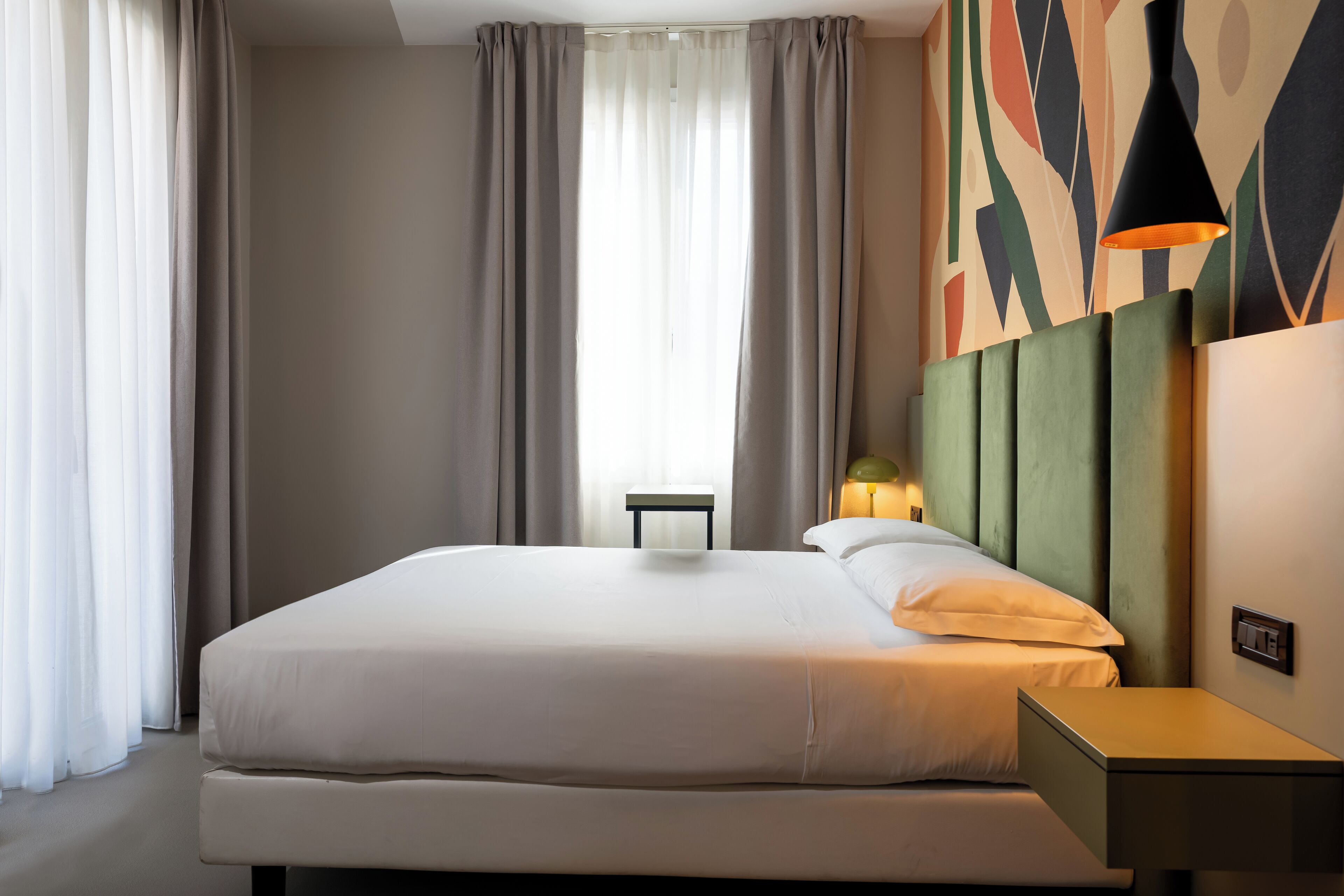 A hotel room with a large bed, a bedside table, a lamp, and a wall with abstract art.