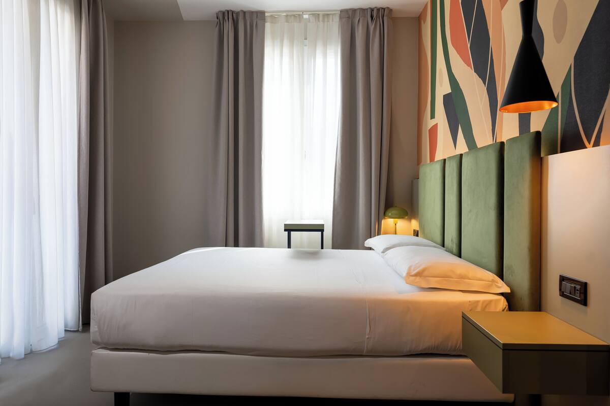 A modern hotel room with a large bed, a bedside table, and a wall-mounted lamp.