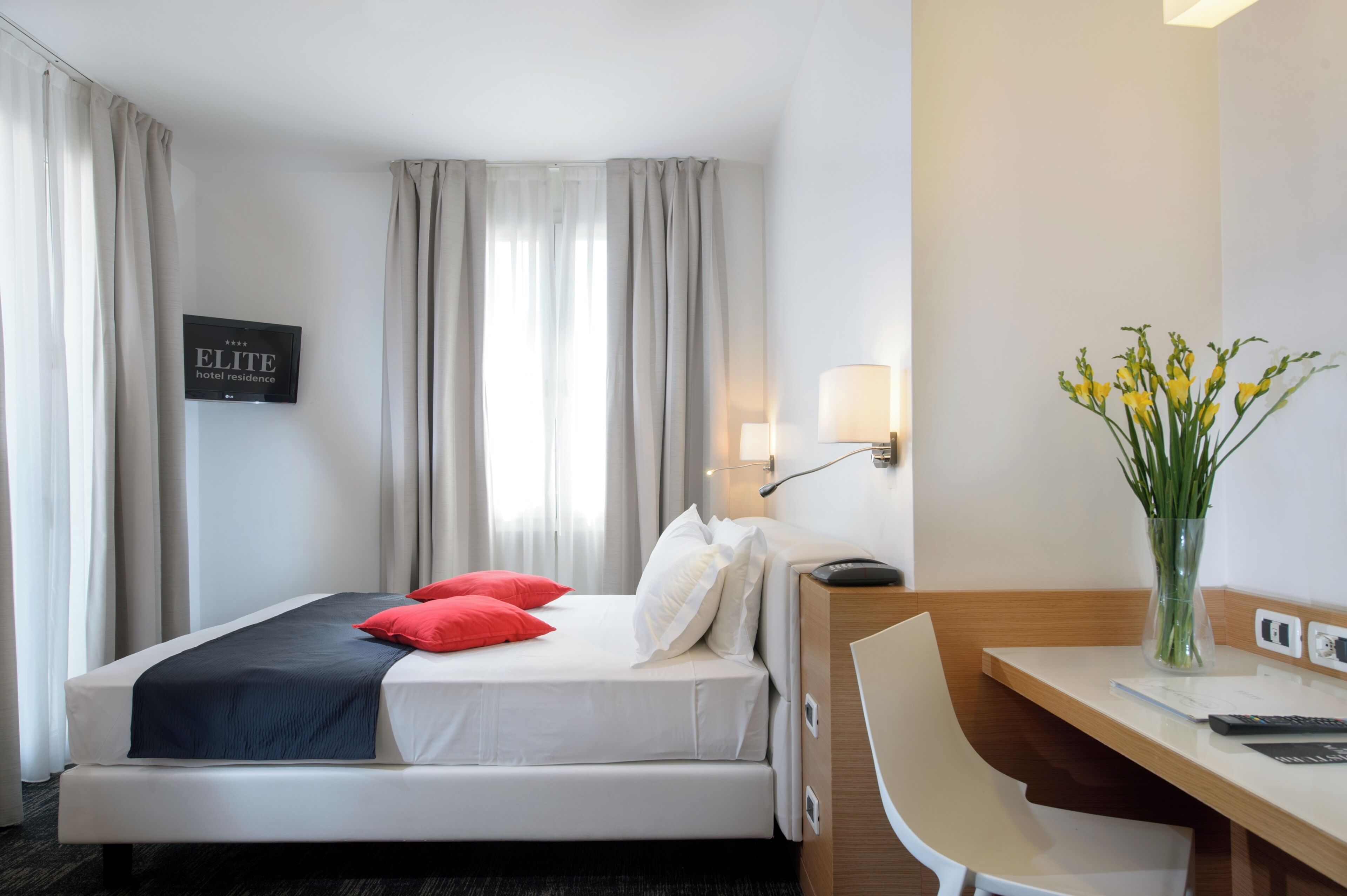 A hotel room with a bed, a desk, a chair, a lamp, and a vase with flowers.