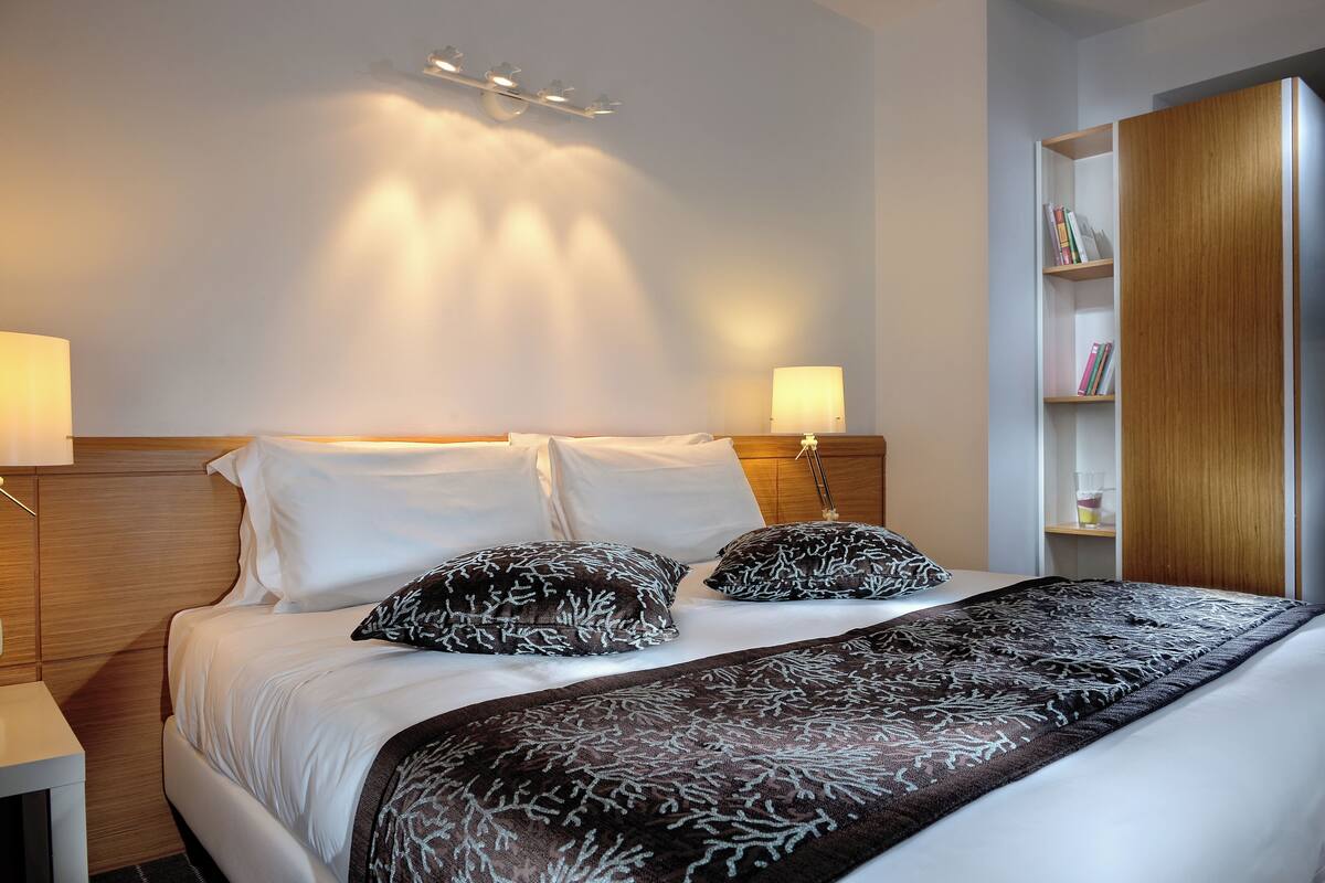 A neatly made bed with white linens and patterned pillows, a bedside lamp, and a wooden headboard.