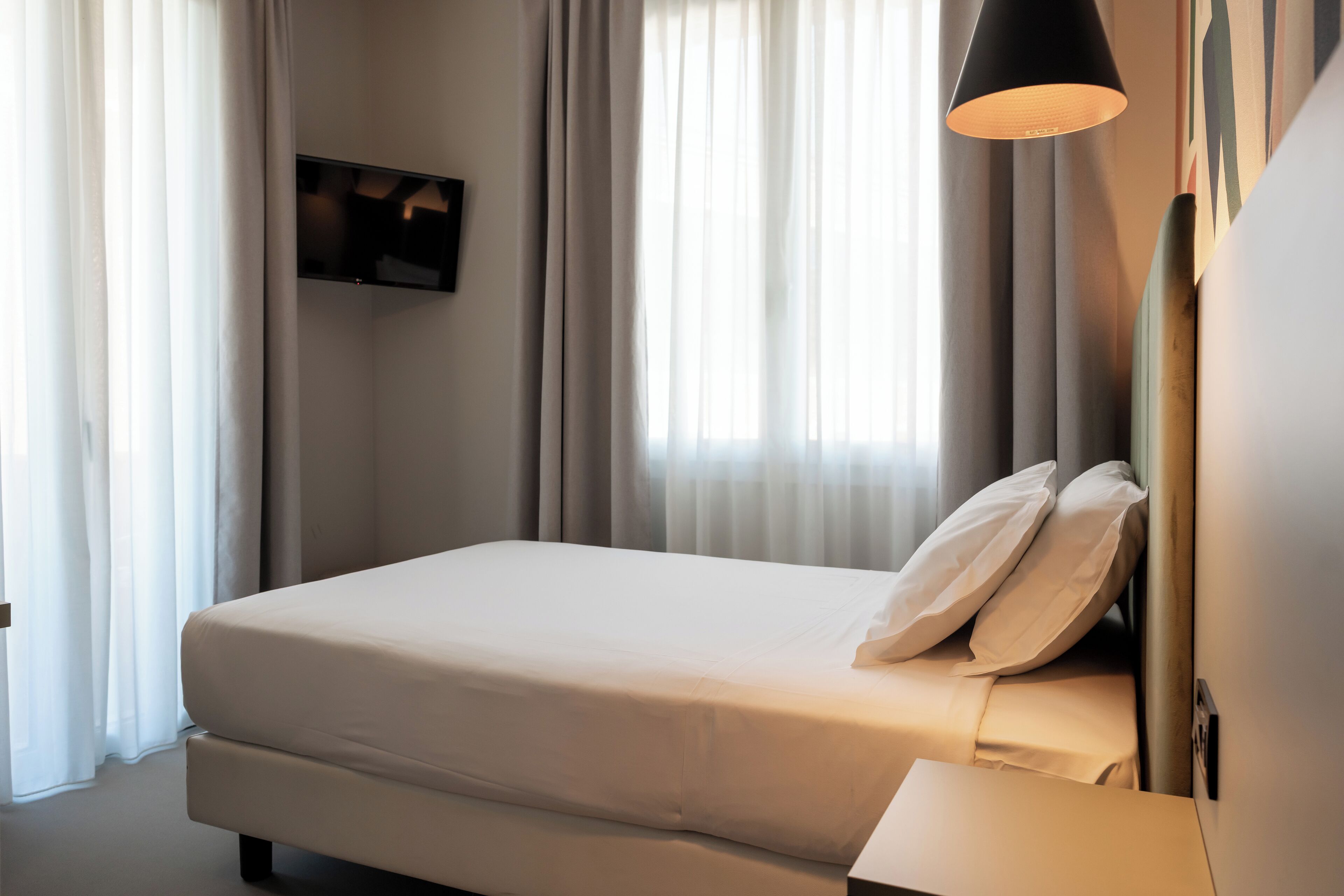 A hotel room with a bed, a bedside table, a TV mounted on the wall, and a window with curtains.