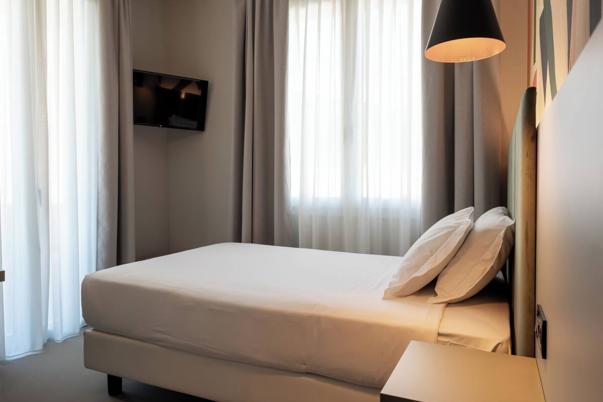 A hotel room with a bed, a bedside table, a flat-screen TV mounted on the wall, and a window with sheer curtains.