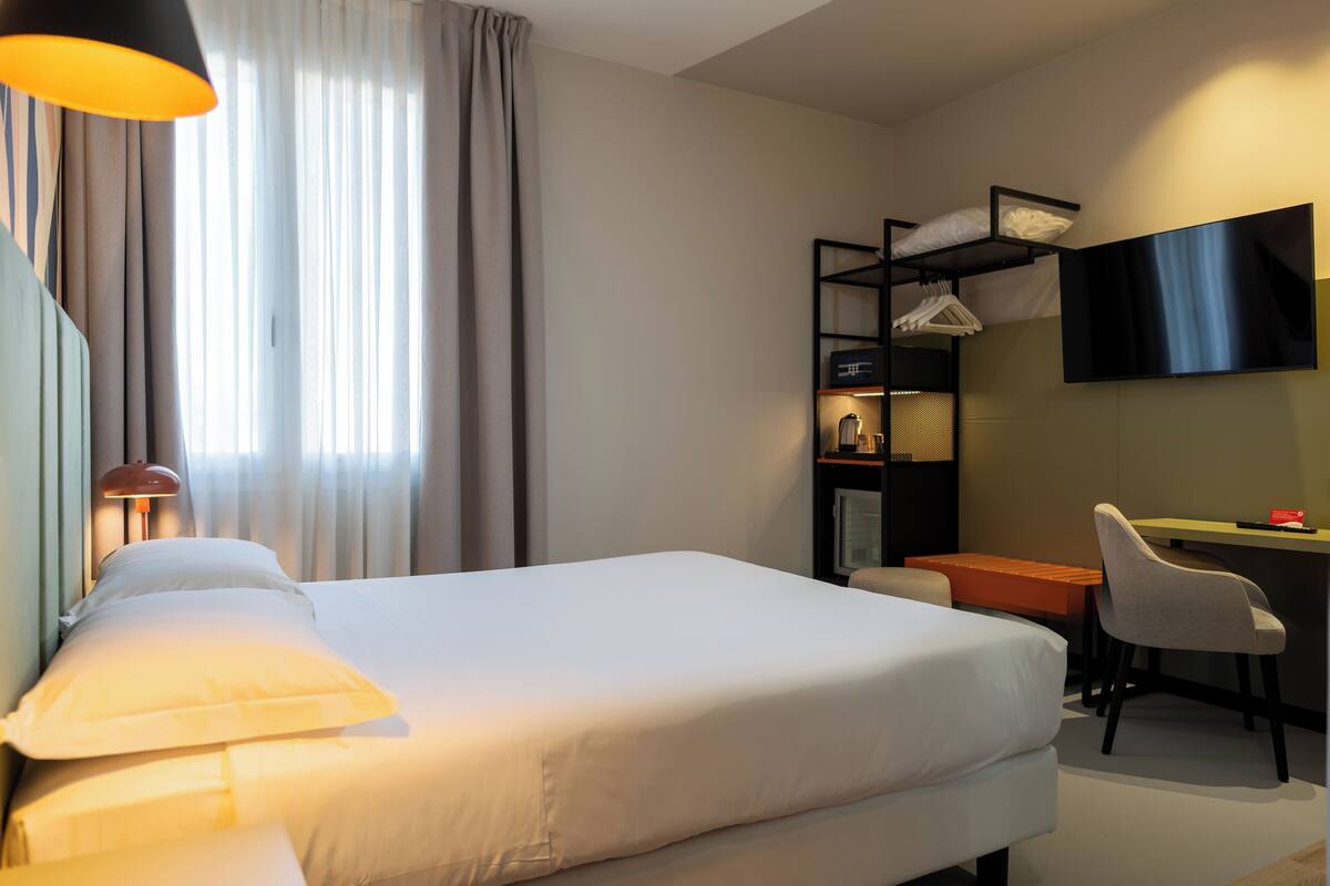 A hotel room with a bed, a desk, a chair, a TV, and a shelf with hanging clothes.