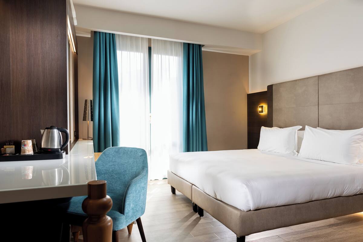 A hotel room with a bed, a desk with a kettle, a chair, and curtains.