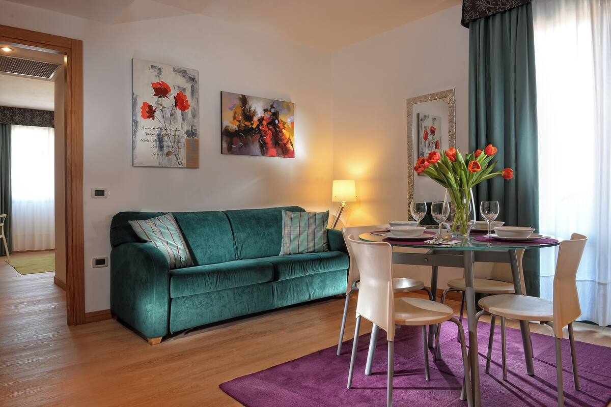 A living room with a green sofa, a dining table with chairs, and a vase of red flowers.