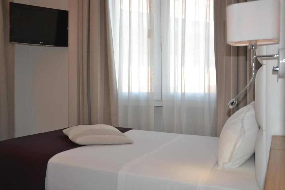 A hotel room with a bed, curtains, a TV mounted on the wall, and a bedside lamp.