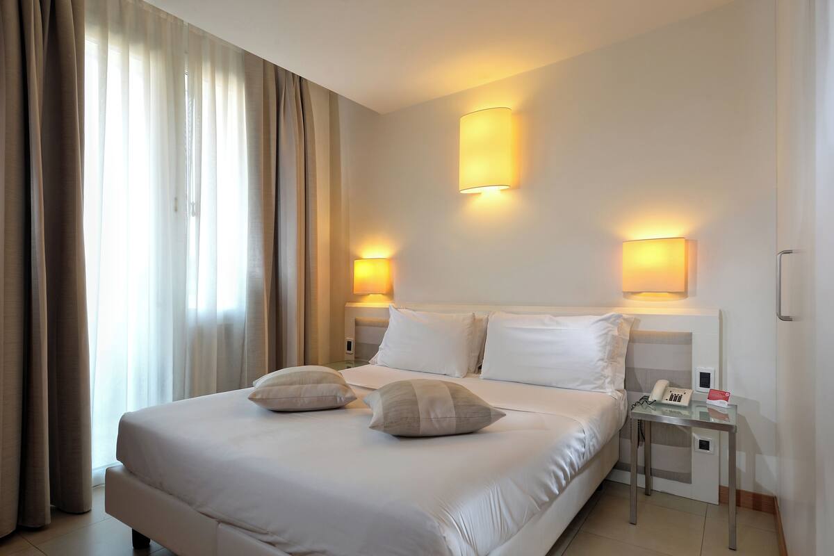 A hotel room with a large bed, two bedside lamps, a bedside table with a phone and a card, and a window with curtains.