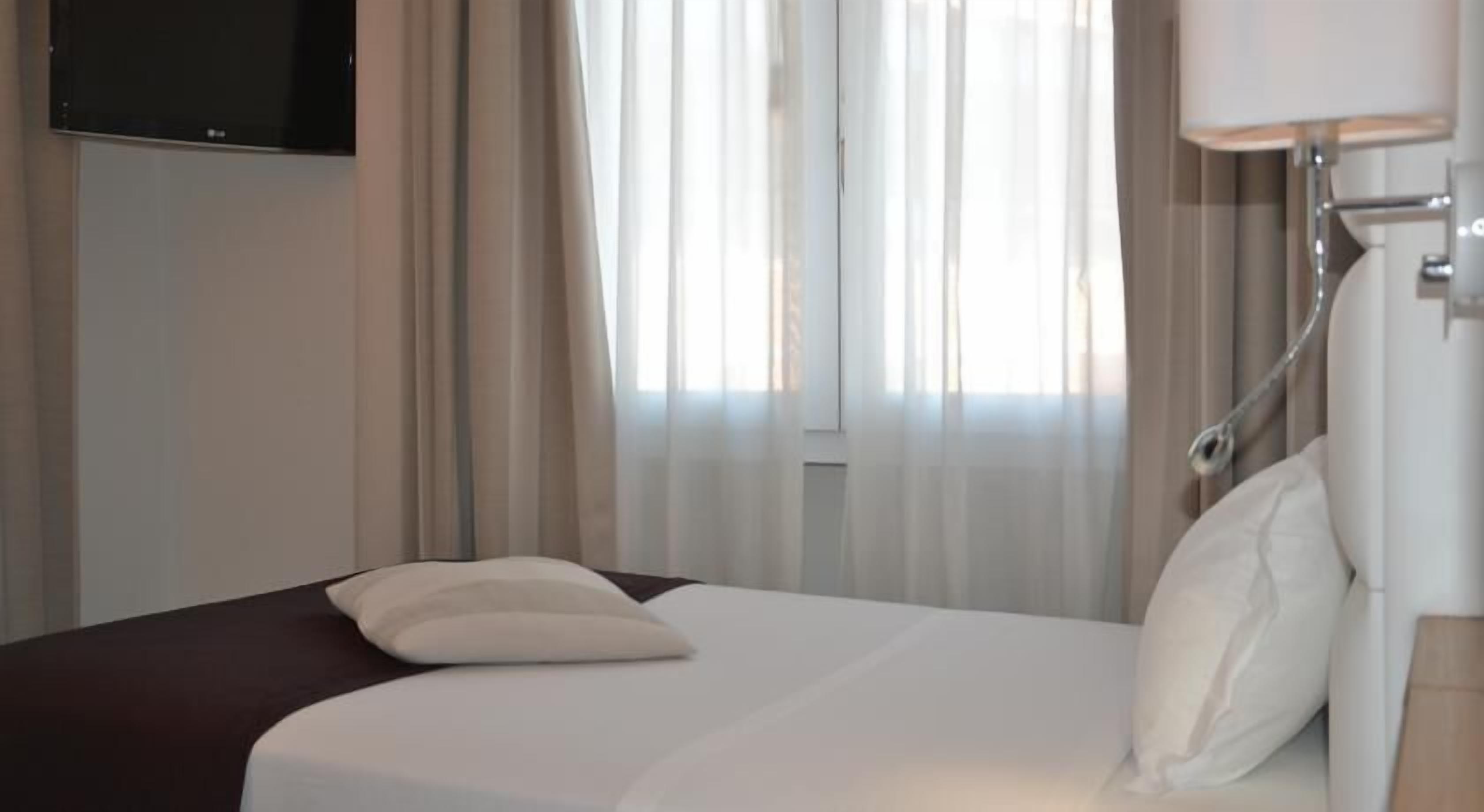 A hotel room with a bed, a TV mounted on the wall, and a window with sheer curtains.