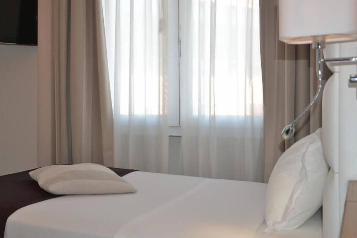 A hotel room with a bed, a TV mounted on the wall, and a window with sheer curtains.