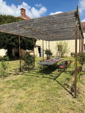 A covered outdoor dining area with a table and chairs, surrounded by grass and small plants. - Le Gîte de Font Valet (Millac)