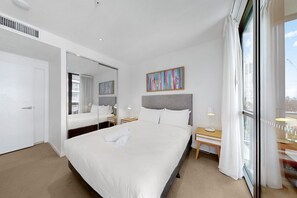 Hotelkamer met een groot bed, nachtkastje met lamp, klein bureau, raam met gordijnen en een kleurrijk abstract schilderij aan de muur.