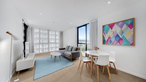 Ein modernes Wohnzimmer mit einer Couch, einem Esstisch mit Stühlen, einem großen Fenster mit Vorhängen und einem farbenfrohen abstrakten Gemälde an der Wand.