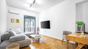 A modern living room with a grey sectional sofa, a wooden coffee table, a TV mounted on the wall, and a dining table with a green lamp. - Modern 2BR in Exarchia | Ensuite Baths & Workspace (Athina)
