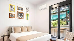 A modern bedroom with a large bed, a small table, and a chair. There are several framed pictures on the wall. - Modern 2BR in Exarchia | Ensuite Baths & Workspace (Athina)