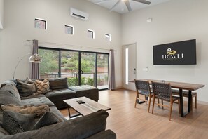 A modern living room with a sectional sofa, a wooden coffee table, and a dining area with a wooden table and chairs. There is a large window with a view of greenery outside. - NEW|Oak Creek Retreat|Townhouse w/Views|FirePit (Sedona)