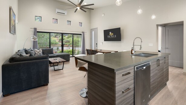 A modern kitchen with a central island, a dining area with a table and chairs, a sofa, and a large window offering a view of greenery. - Moondance @ Oak Creek | Townhouse w/Views| FirePit (Sedona)