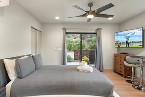 A bedroom with a bed, a ceiling fan, a TV, a dresser, and a view of a garden. - Cimaron Butte Studio | Kitchenette & Laundry (Sedona)
