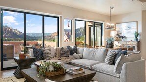 A modern living room with a large sectional sofa, a coffee table with decorative items, and a view of the mountains through large windows. - Sycamore Canyon Luxe Retreat | 180° Views + Hottub (Sedona)