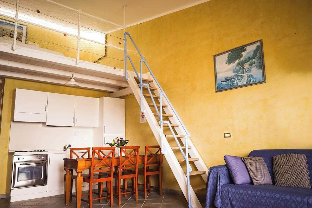 Un ambiente compatto con angolo cottura, zona pranzo e una scala che conduce a un piano superiore.