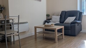 Ein Wohnzimmer mit Holzboden, einer grauen Couch mit Kissen, einem kleinen Holztisch, einem Metallstuhl, einem Gemälde an der Wand und einer Uhr.