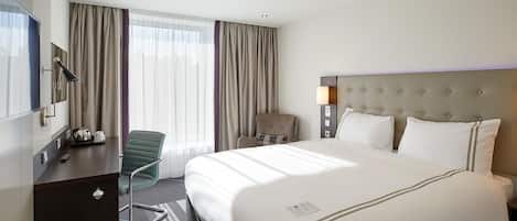 Una habitación de hotel con cama, escritorio, silla, lámpara y ventana con cortinas.