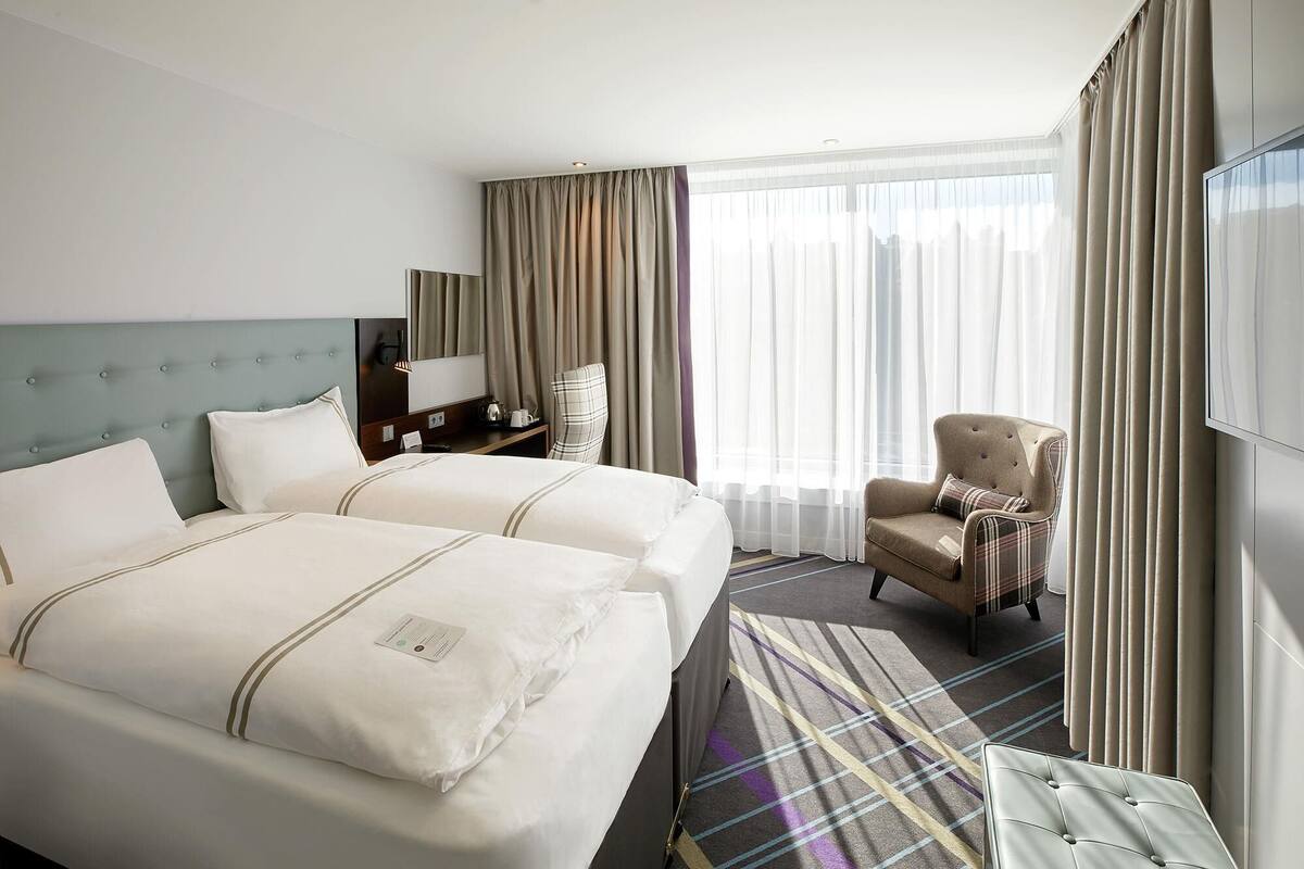 A hotel room with two beds, a chair, a TV, and a large window with curtains.