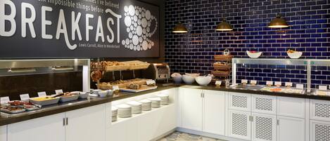 A breakfast buffet with various food items, a wall with a quote, and a blue-tiled backsplash.
