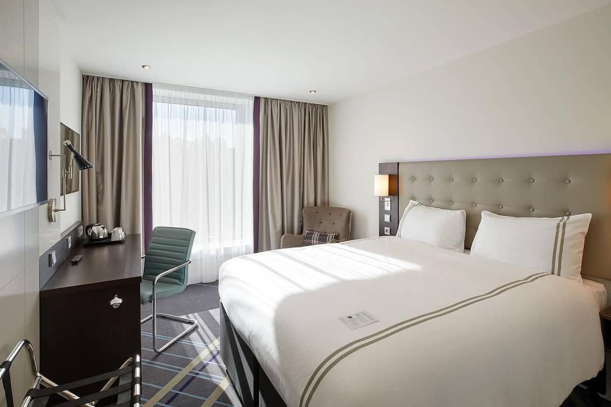 A hotel room with a bed, a desk, a chair, a lamp, and a window with curtains.