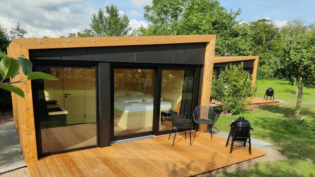 A modern, compact cabin with a wooden deck, large sliding glass doors, and a small outdoor seating area. - Cabin in Nature near Scenic Trails (Scherpenzeel (FR))