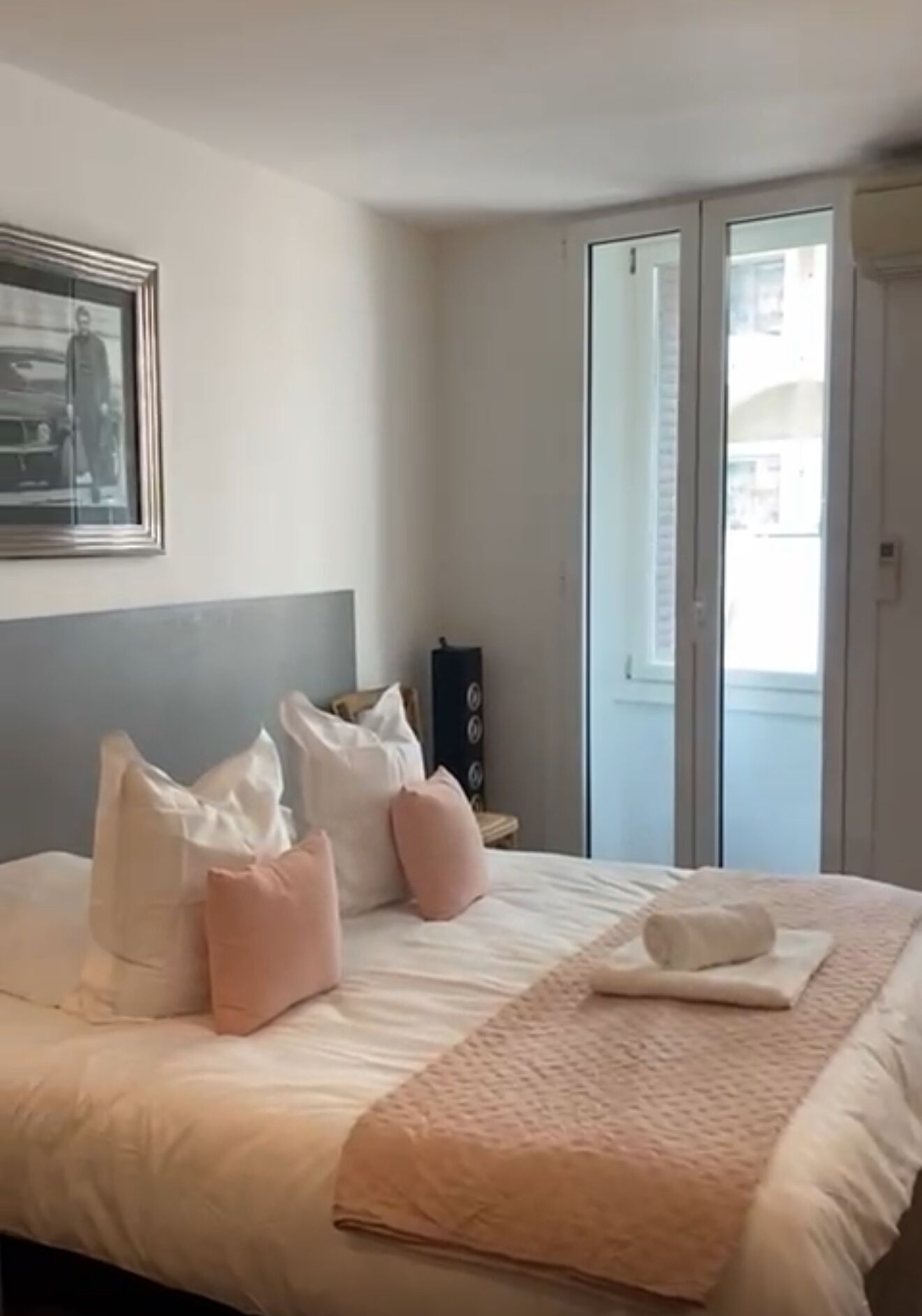 A hotel room with a bed, pillows, a bedside table, a lamp, and a window.
