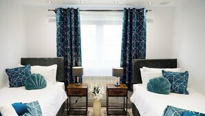 A modern bedroom with two beds, matching blue and white bedding, and a vase of white flowers on the floor.