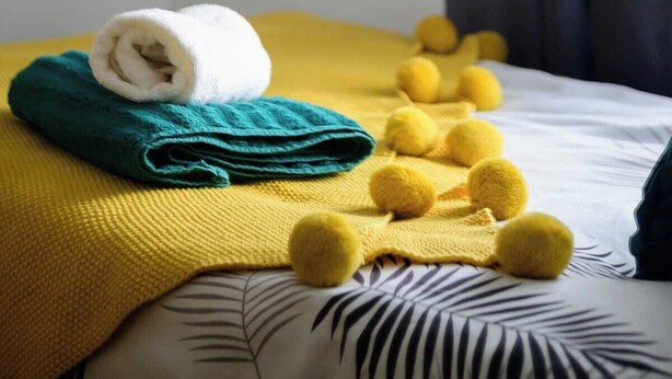 A neatly made bed with a rolled white towel, a teal towel, and a yellow blanket with pom-poms.