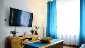 A room with a flat-screen TV mounted on the wall, a wooden cabinet with a plant, a table with a fruit bowl, and a window with blue curtains.