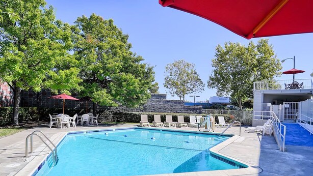 Una piscina rectangular con un trampolín, rodeada de sillas de descanso y sombrillas, con árboles y un edificio al fondo.