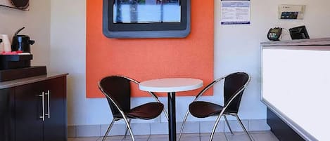 Een klein eetgedeelte met een ronde tafel en twee stoelen, een aan de muur bevestigde televisie, en een toonbank met een kassa.