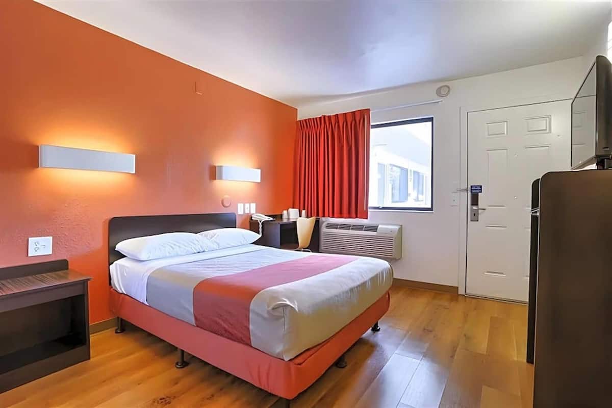 Habitación de hotel con cama, mesita de noche, luz empotrada en la pared, ventana con cortinas y una puerta.