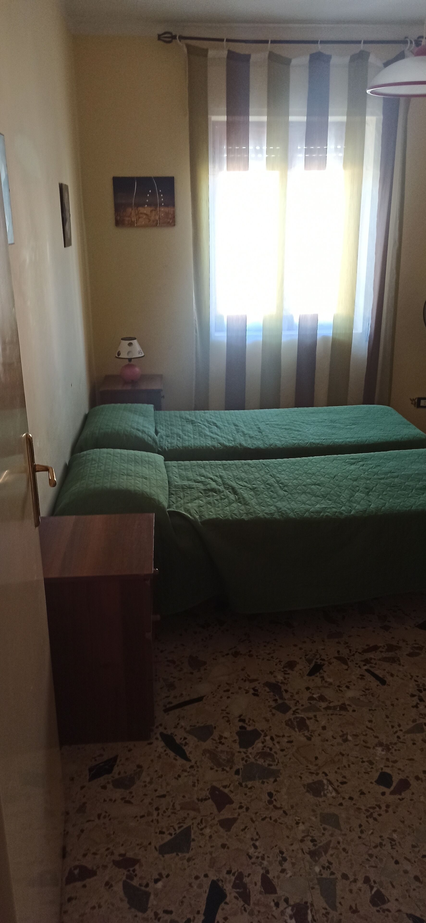 Een kamer met een bed, een klein tafeltje, een lamp en een schilderij aan de muur.